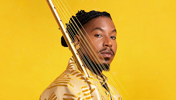 Headshot of Chief Adjuah. He wears a gold shirt against a yellow background and holds a stringed instrument. 