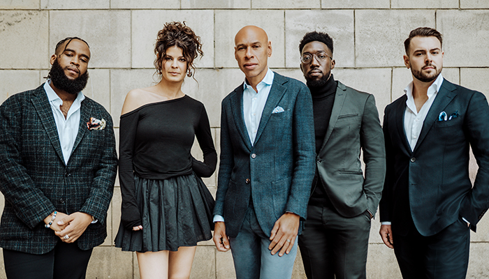 Four men and a woman stand against a stone wall, wearing suits and a dress. Joshua Redman stands in the center.