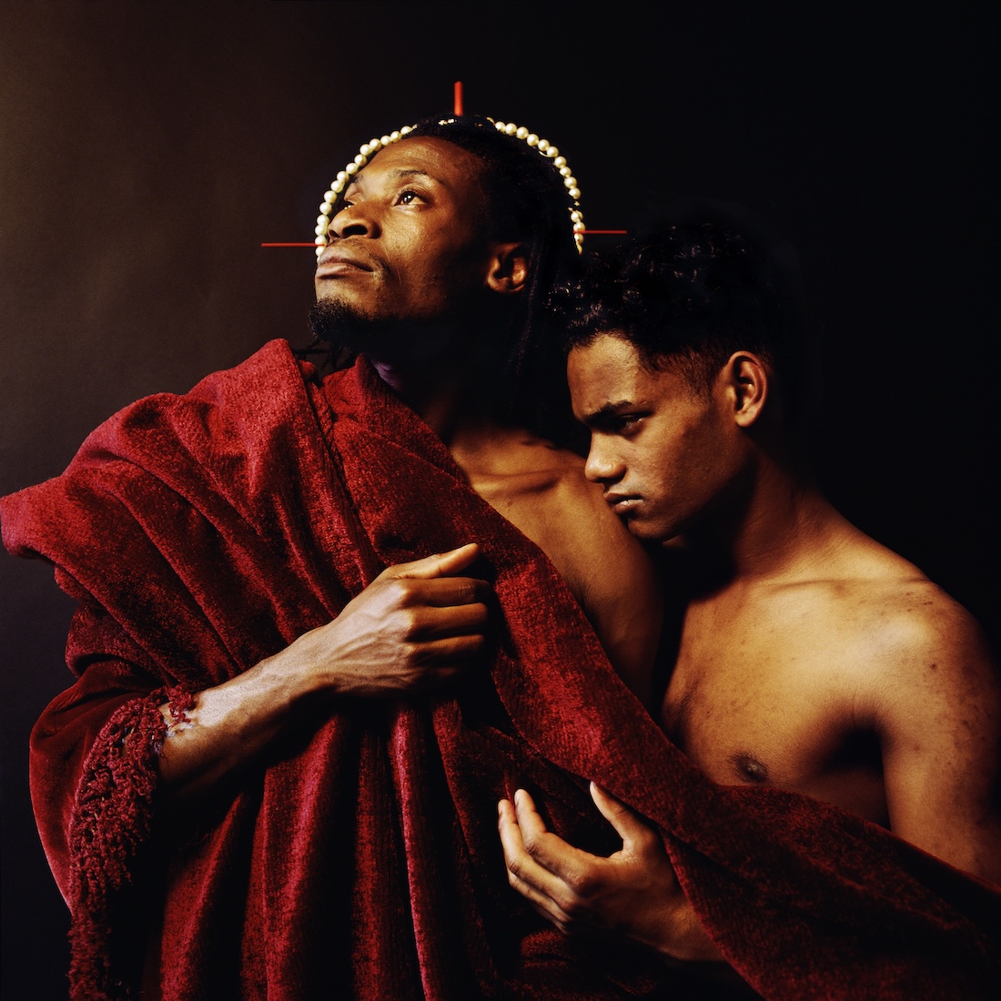 A black man wearing a strung pearl halo and a red shawl looks upward as a younger Black man stands close by his side and leans on him.