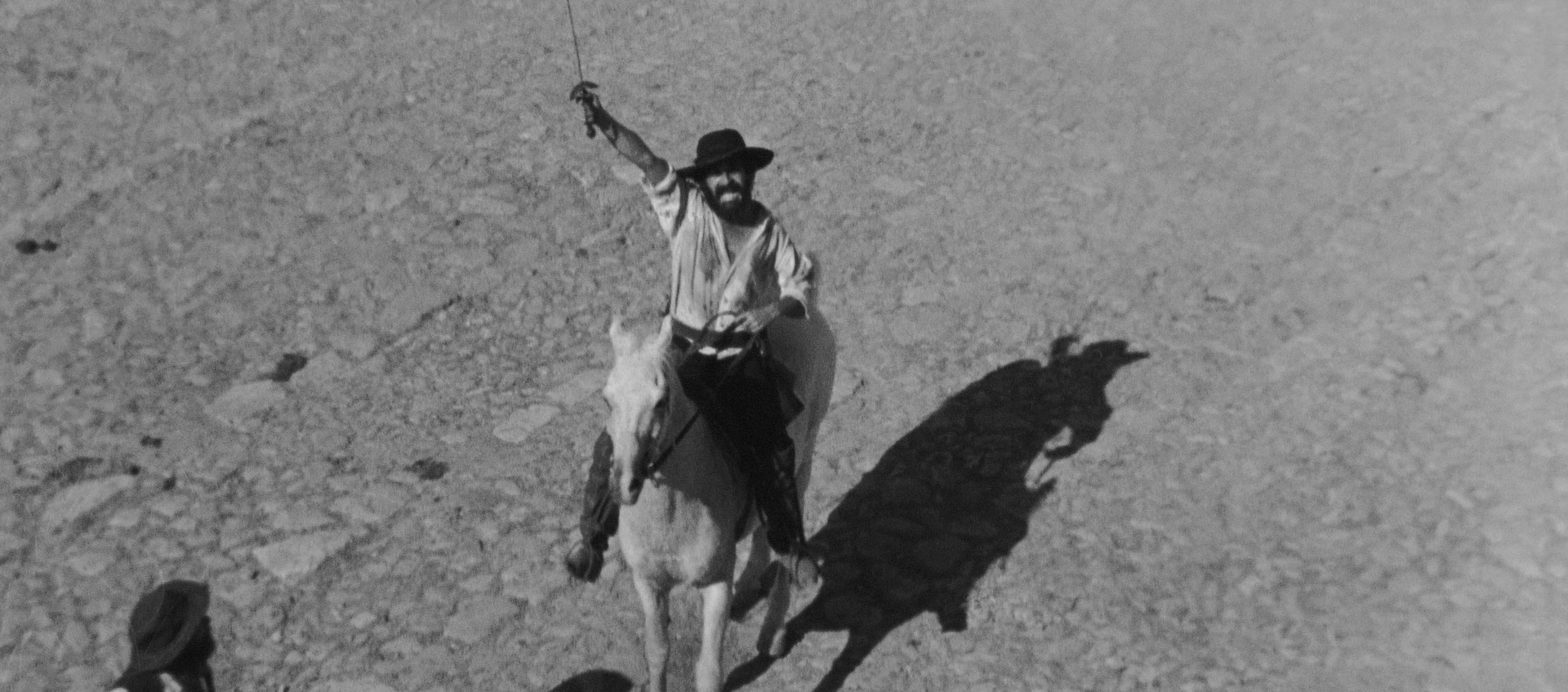 Black and white film still of a man sitting on a horse in the desert and holding a sword upward.