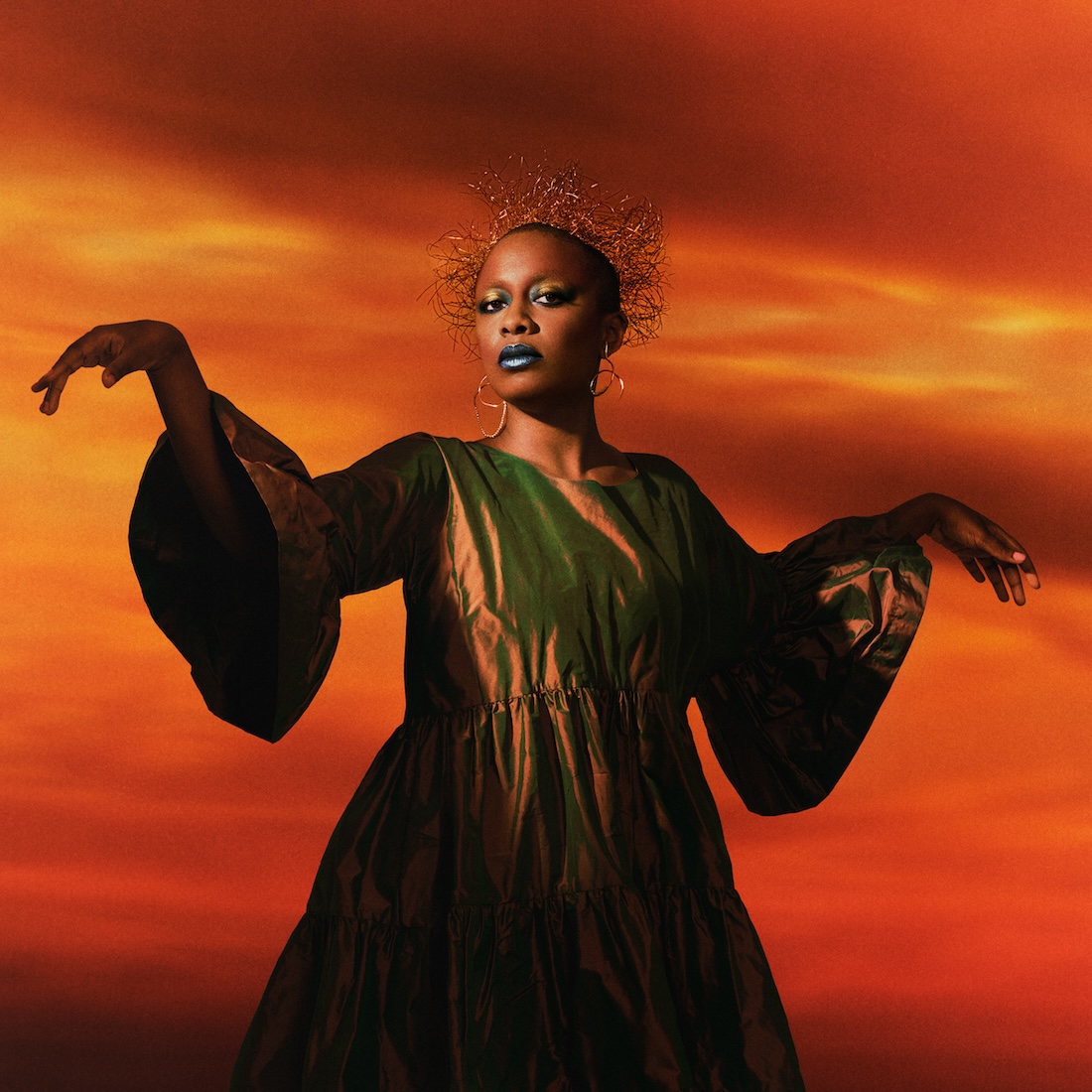 A Black woman stands in front of a fiery orange background, wearing a wiry headdress and a metallic bronze gown, her arms partially outstretched.