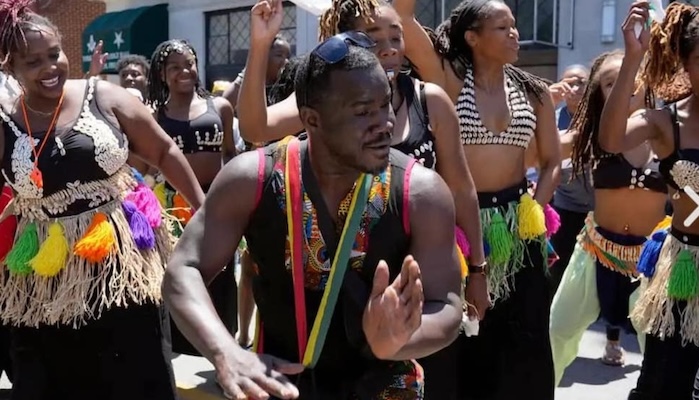 A group of young Black men and women in colorful costumes dance in the street.