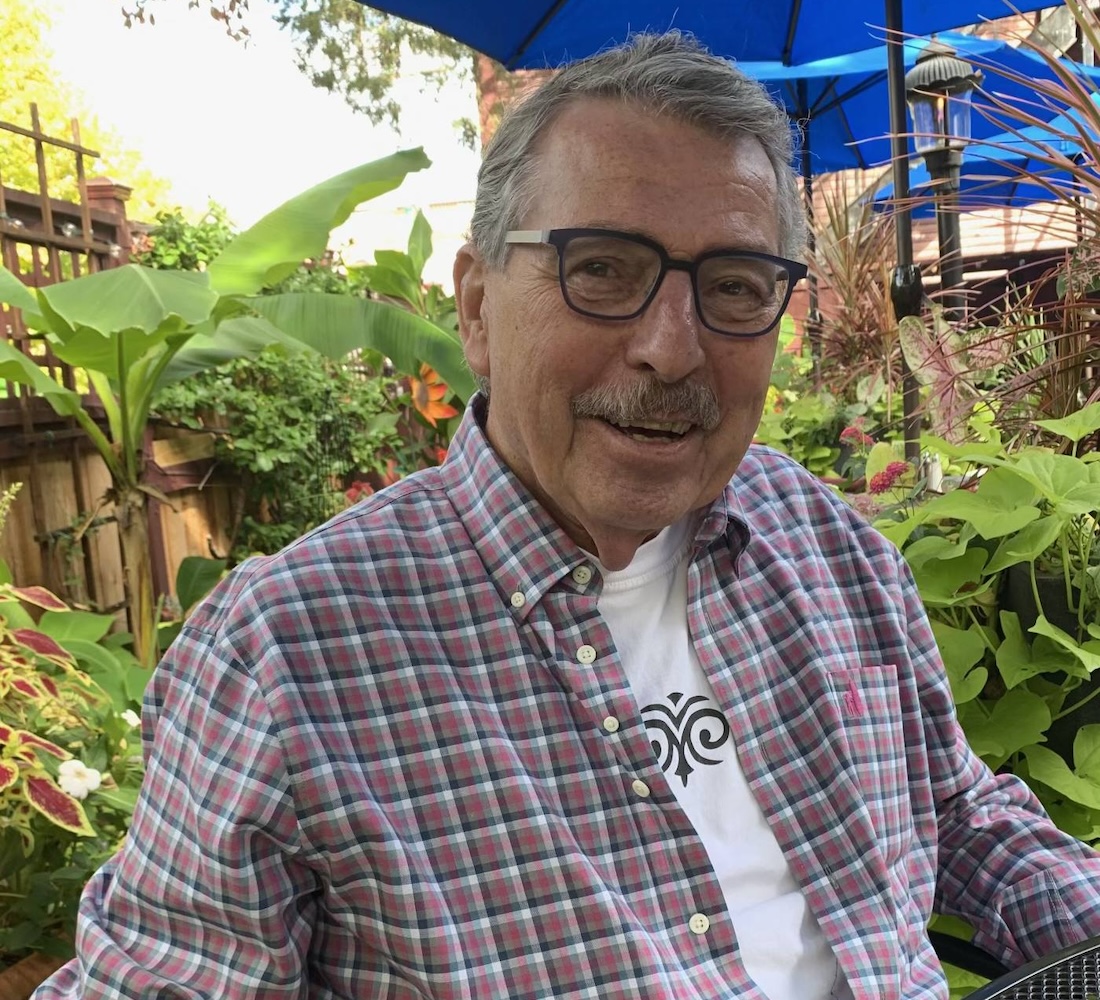 A man with short gray hear and glasses smiles for the camera as he sits in a plant-lined patio.