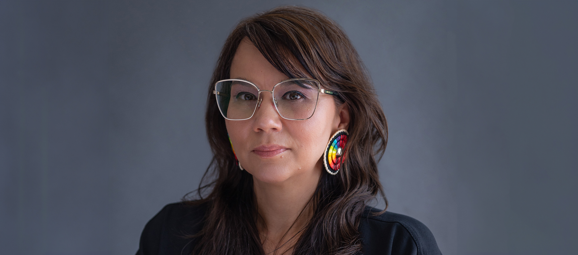 POPortrait of Tanya Lukin Linklater. She has long brown hair, a medium skin tone, and wears geometric silver glasses, beaded earrings, and a dark shirt.