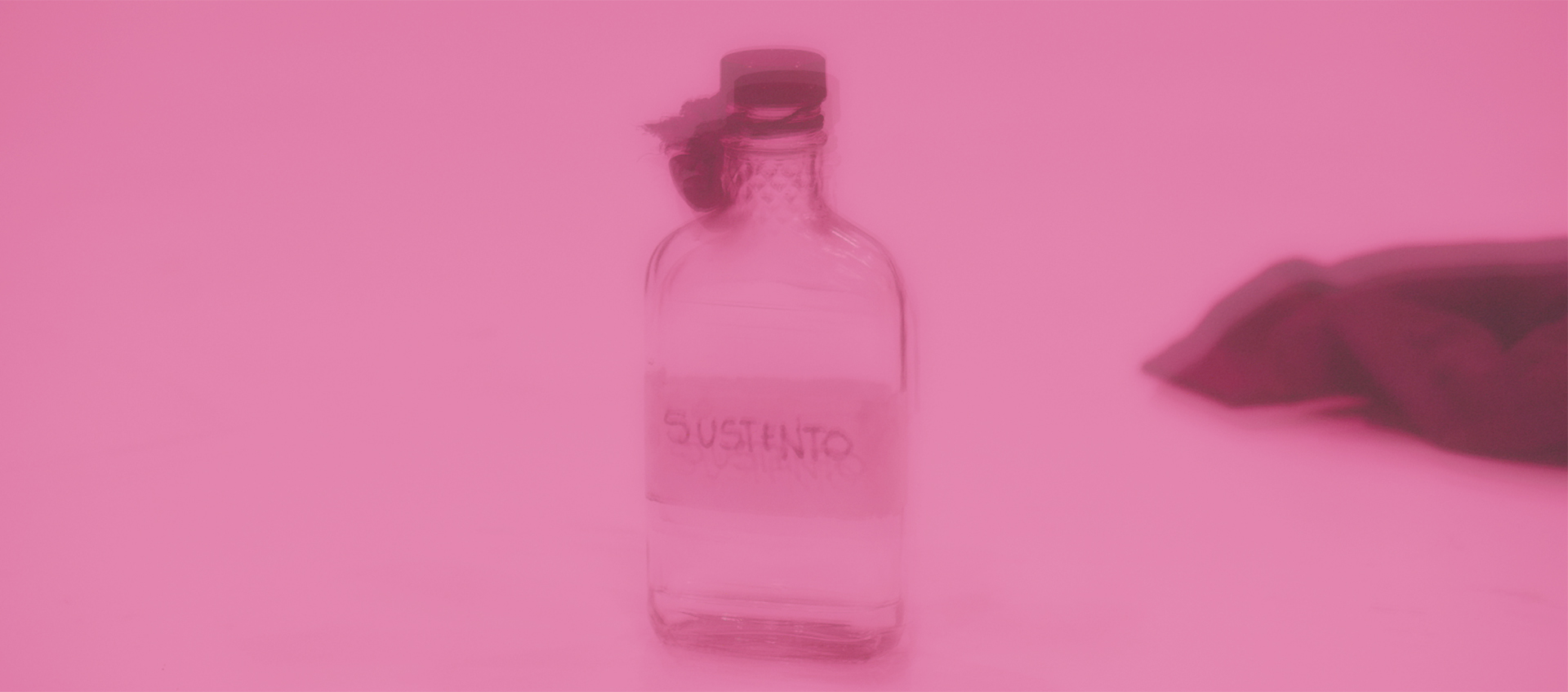 A blurry, pink-tinged image of a clear, lidded sloped shoulder, flattened glass bottle labeled with the word susteno.