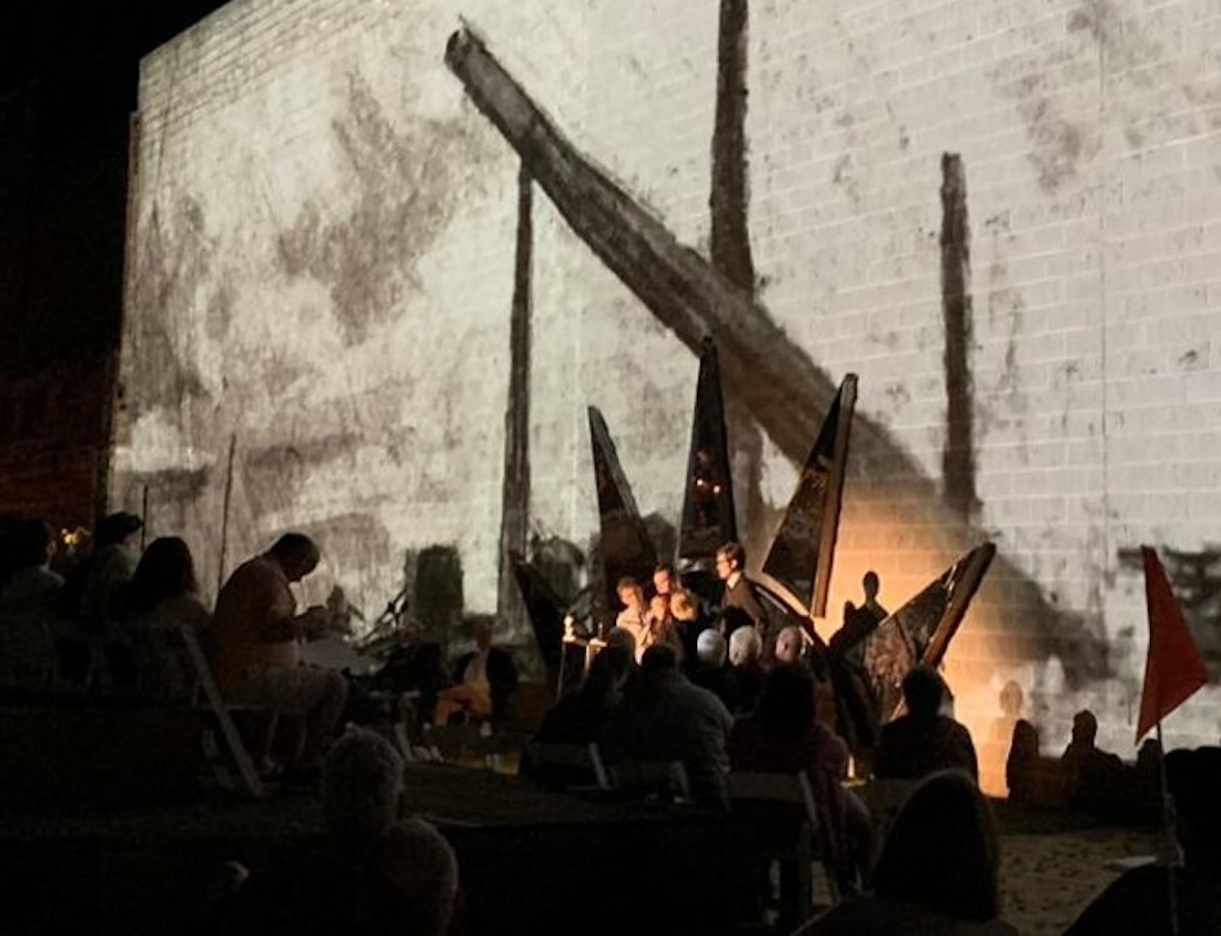 An outdoor performance with a group of people gathering on a stage with an image of a charcoal drawing projected large behind them.