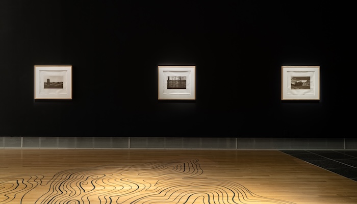 A dark wall in a gallery holds a row of three framed photographs over a lit wooden floor bearing topographical lines.