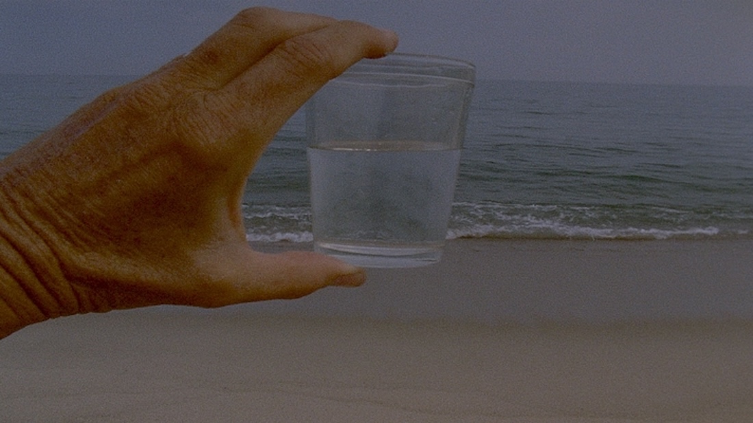 A grainy color image of a woman’s hand holding a drinking glass up in front of her view of a sandy beach and the ocean.