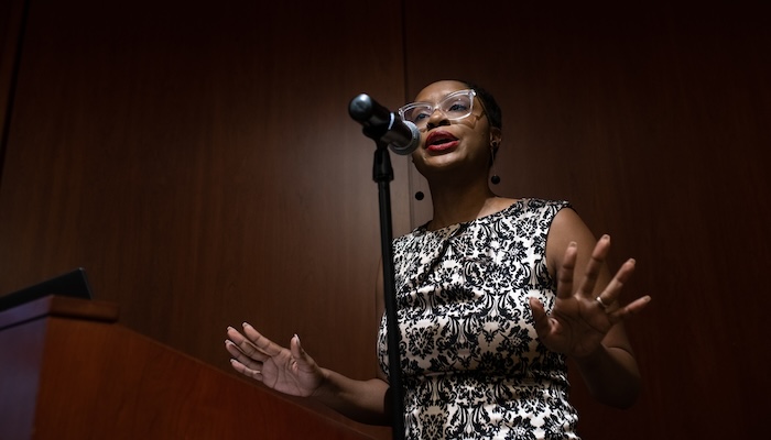 A Black woman speaks into a microphone, her hands stretched out in front of her.