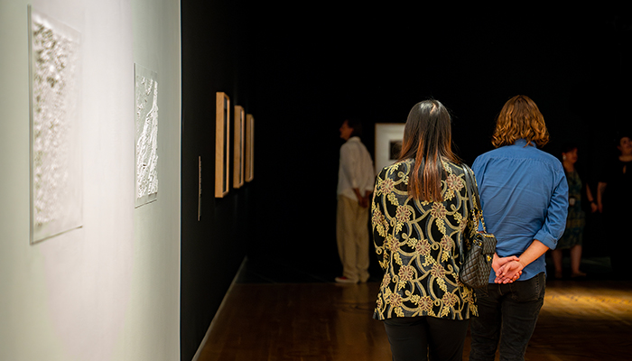 Two people walk out of a white gallery space displaying white topographical maps and into a dark gallery with framed photographs.