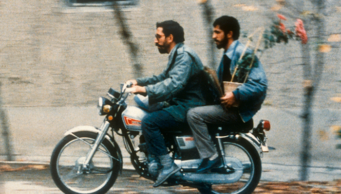 Two bearded men are riding on a motorcycle together wearing blue jackets. The man in front is wearing sunglasses.  