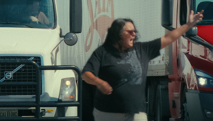 A qoman stands next to a semi truck with her hand in the air. She appears to be yelling.