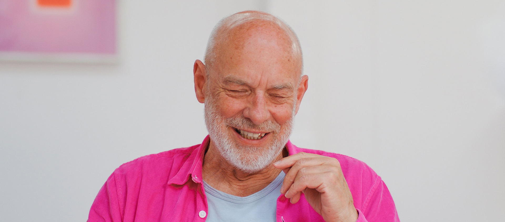 An older bald man with a white beard, wearing a pink shirt, looks downward and smiles.