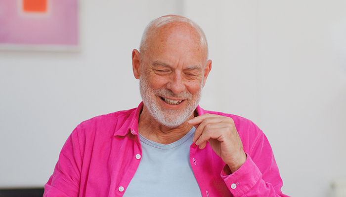 Close-up of a bald man with a white beard. He is wearing a pink button shirt. The background is a white wall.  