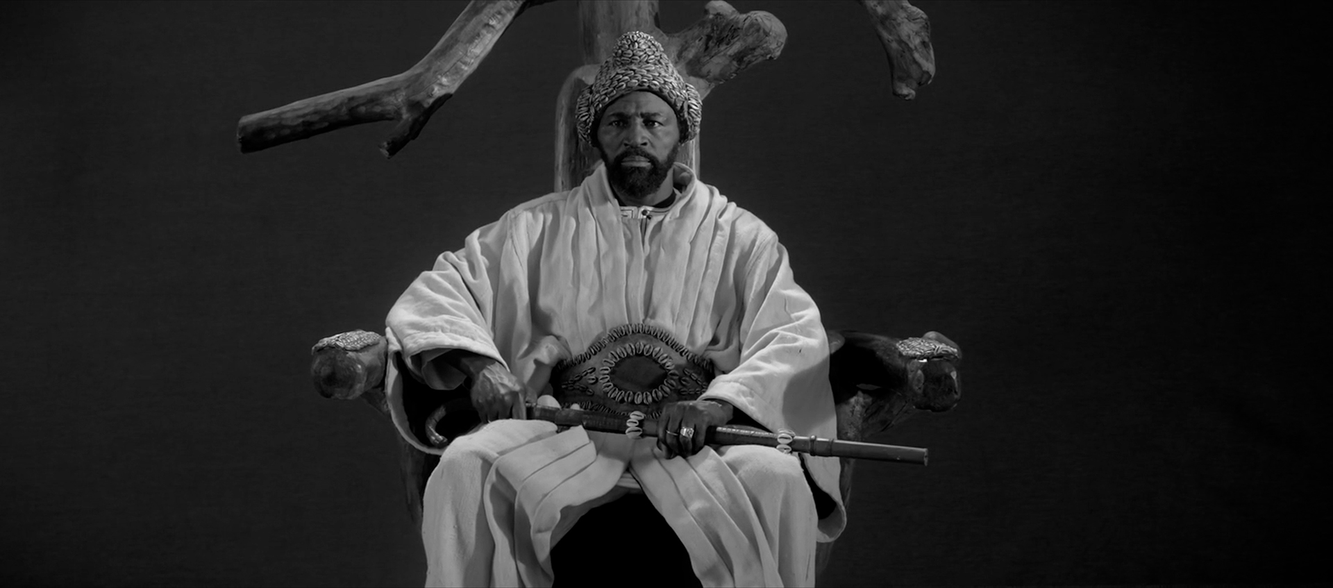 A black-and-white image of a man sitting on a throne with a rifle across this lap. He is wearing a knit hat and a white robe. 