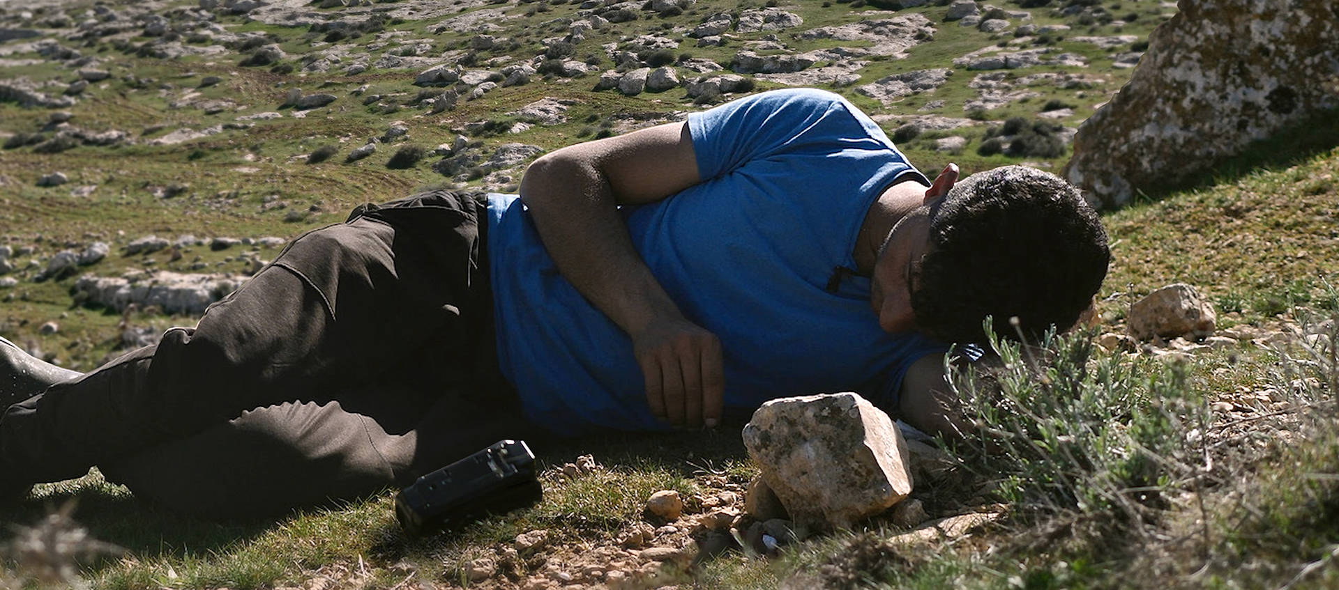 a young man lays on the ground amongst rocks, a camcorder is next to him.