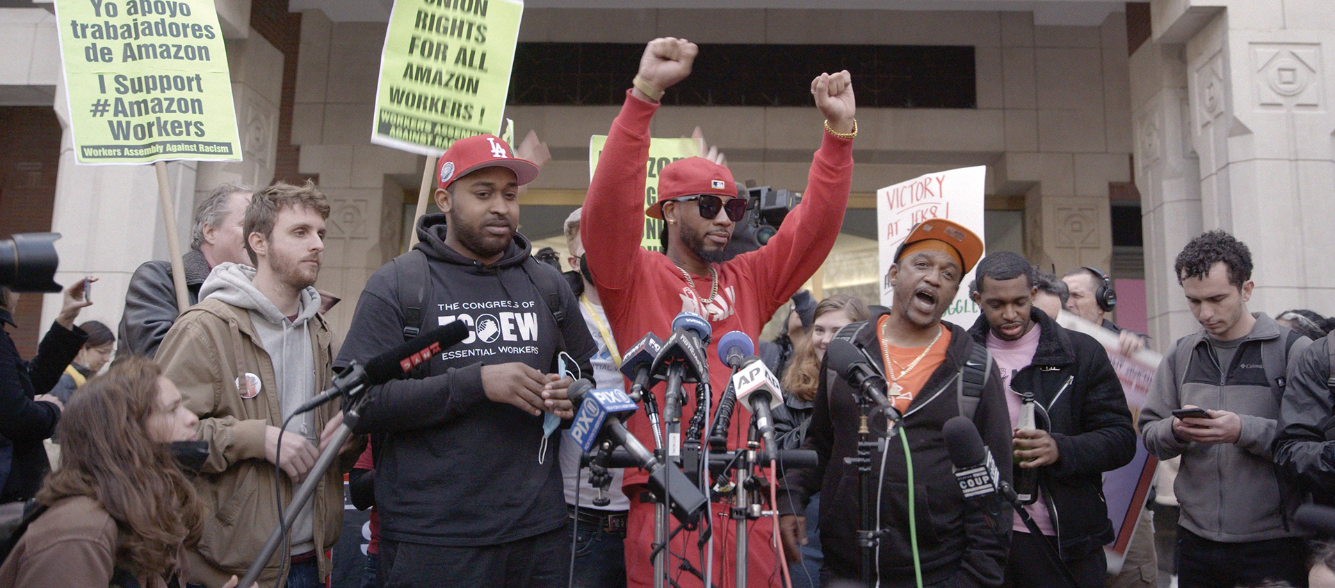 Individuals standing in front of news microphones, the man in the center has his fists raised above his head.