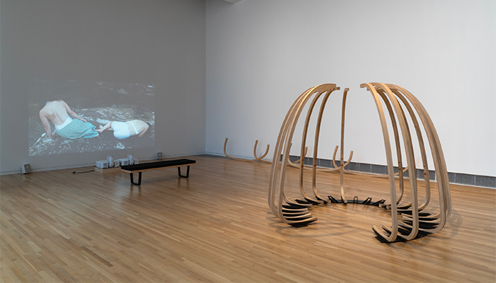 A tall, semicircular sculpture of fifteen curved wooden ribs on a metal base sits near a video projection depicting two figures reclining on a rock. 