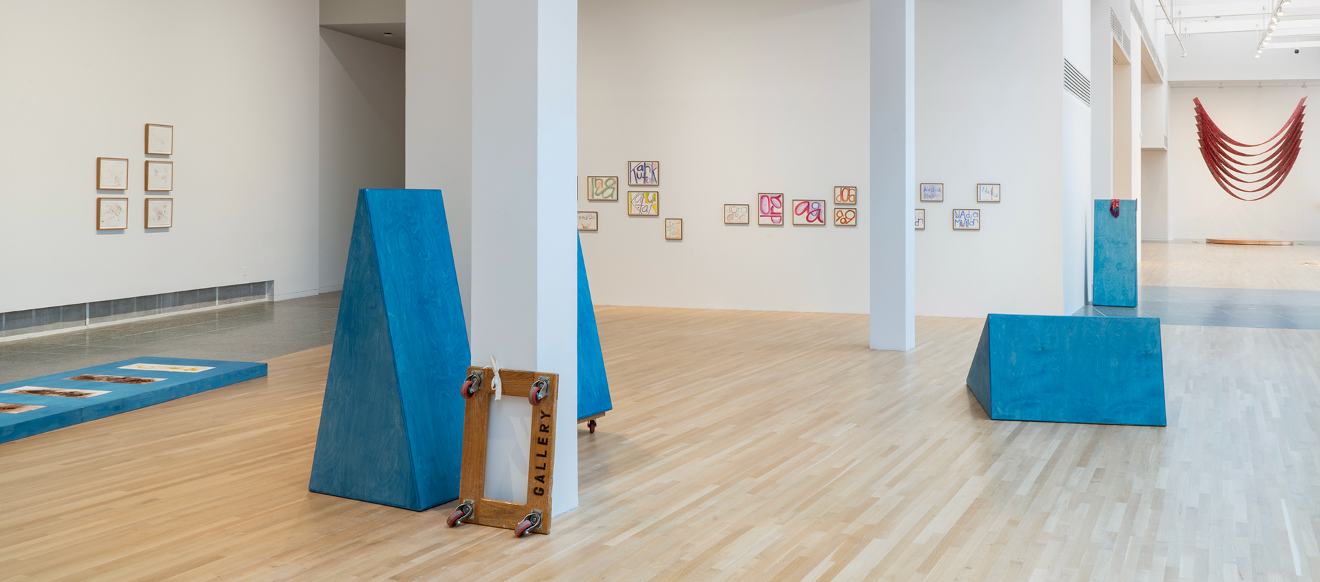 Gallery view of four freestanding triangular blue sculptures, prints displayed on a riser and the walls, and a hanging, U-shaped, red fabric work.