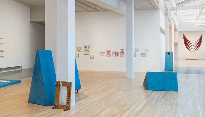 Gallery view of four freestanding triangular blue sculptures, prints displayed on a riser and the walls, and a hanging, U-shaped, red fabric work.