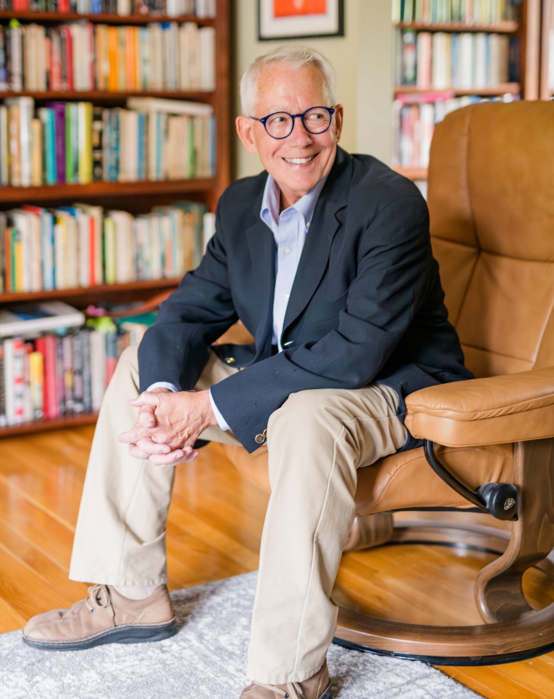 A man with white hair and glasses sits in a book-lined space and smiles as he looks to his left.