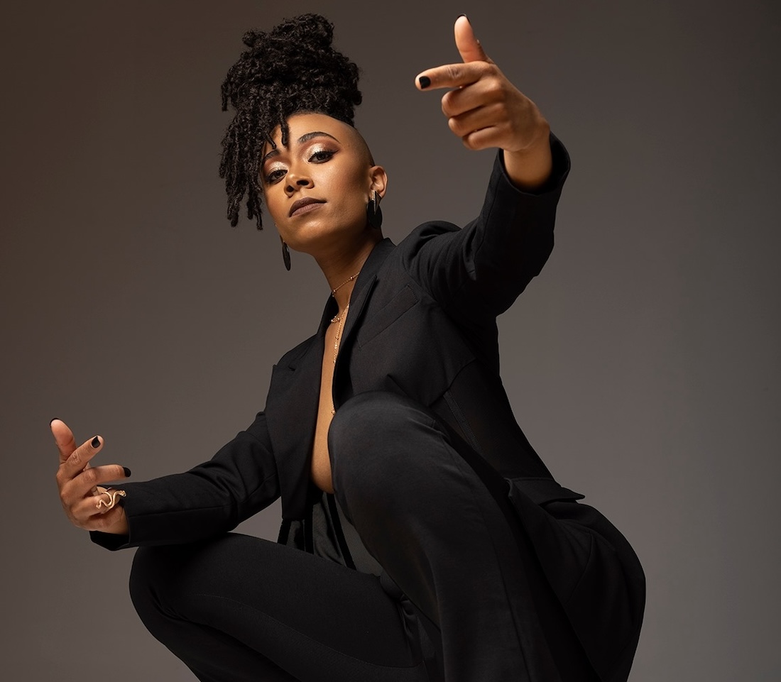 A Black woman wearing a black suit and a braided updo squats with arms extended as she looks down toward the camera.