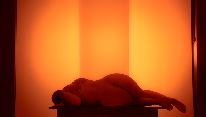 The artist lies naked on a table against a backlit amber background. She is on her side, facing away from the camera, with her head turned downward.