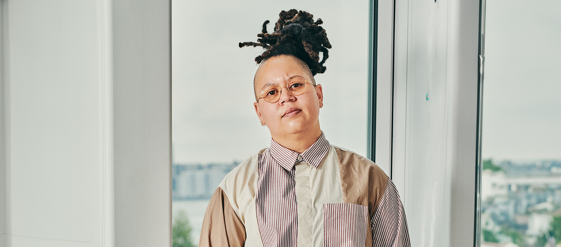 Meg Onli wears wire-rimmed glasses and a collared shirt made from multiple fabrics. Her locs are styled in a topknot.