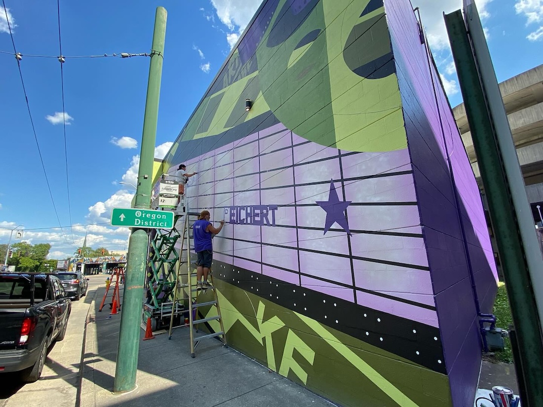 Two artists paint a mural on an exterior wall of a movie theater.