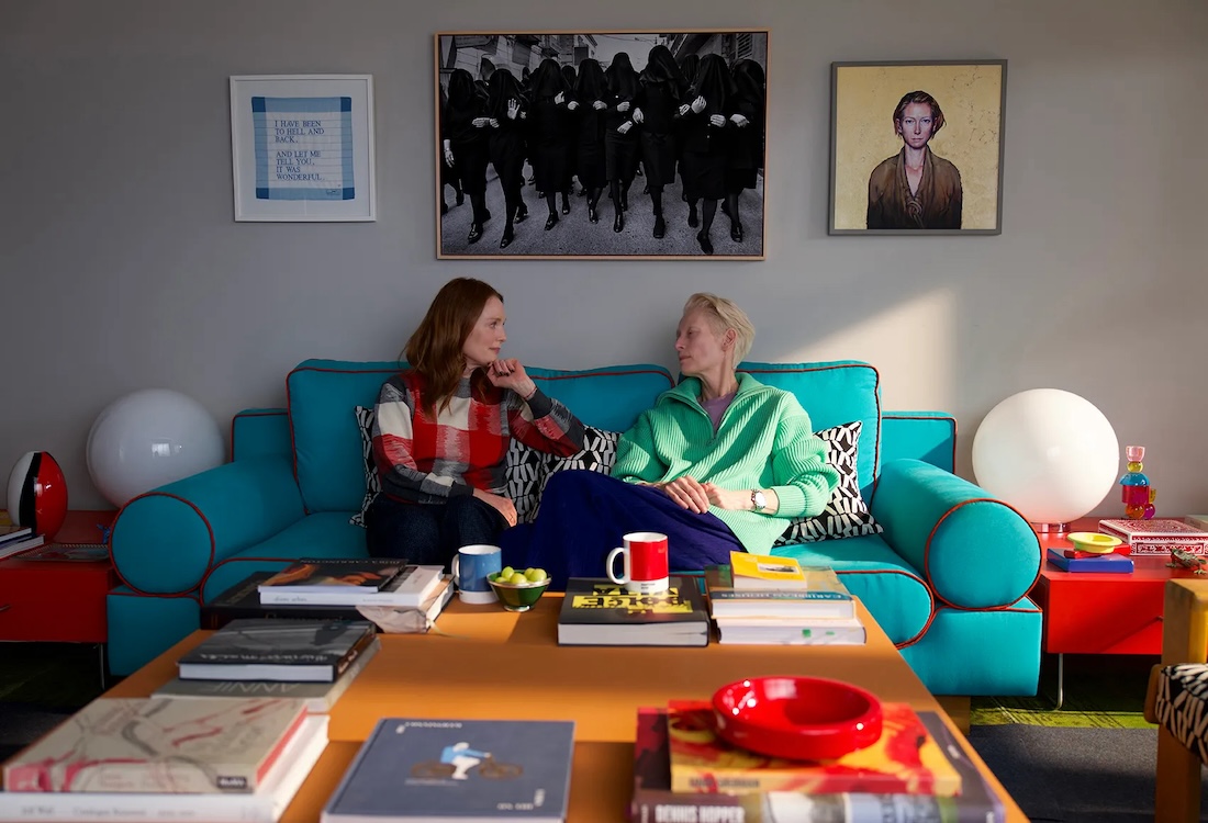 Two women sit facing each other on a blue couch in a colorful living room with art on the walls.