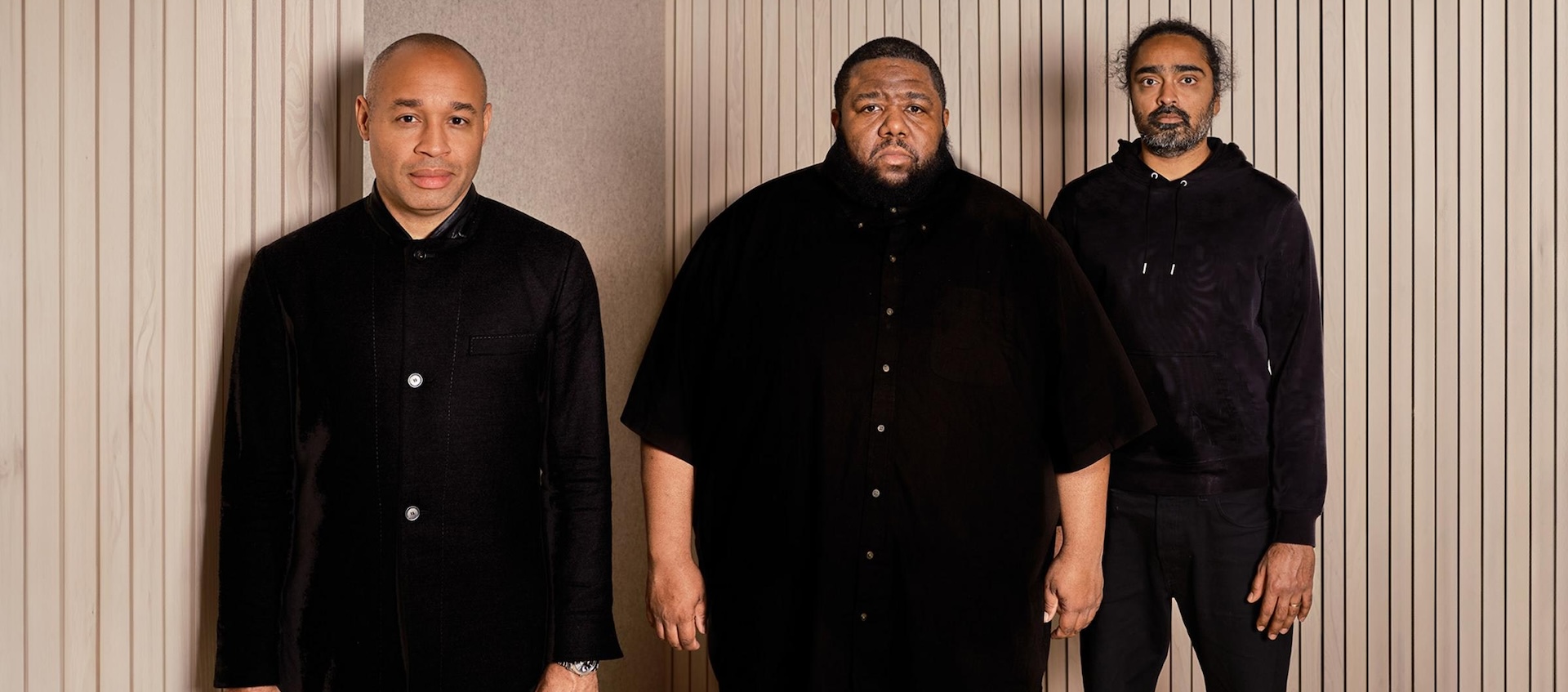 Three men in black stand in a space with beige, vertically-lined walls.