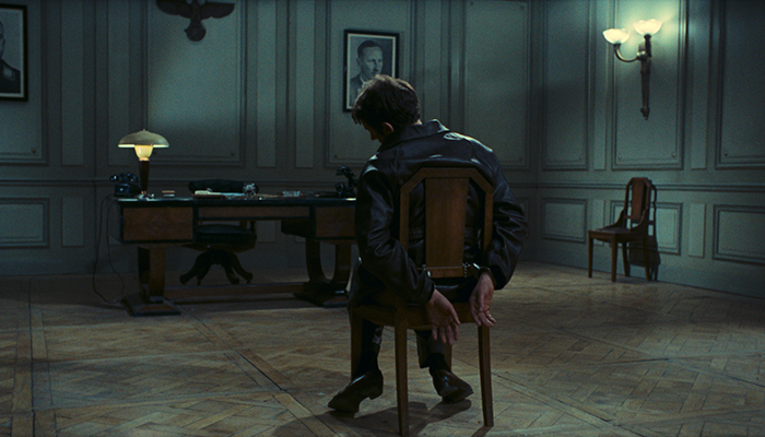 A man is tied to a chair in the middle of a large room. There is a desk in front of him and two photos of military officers on the otherwise empty walls.