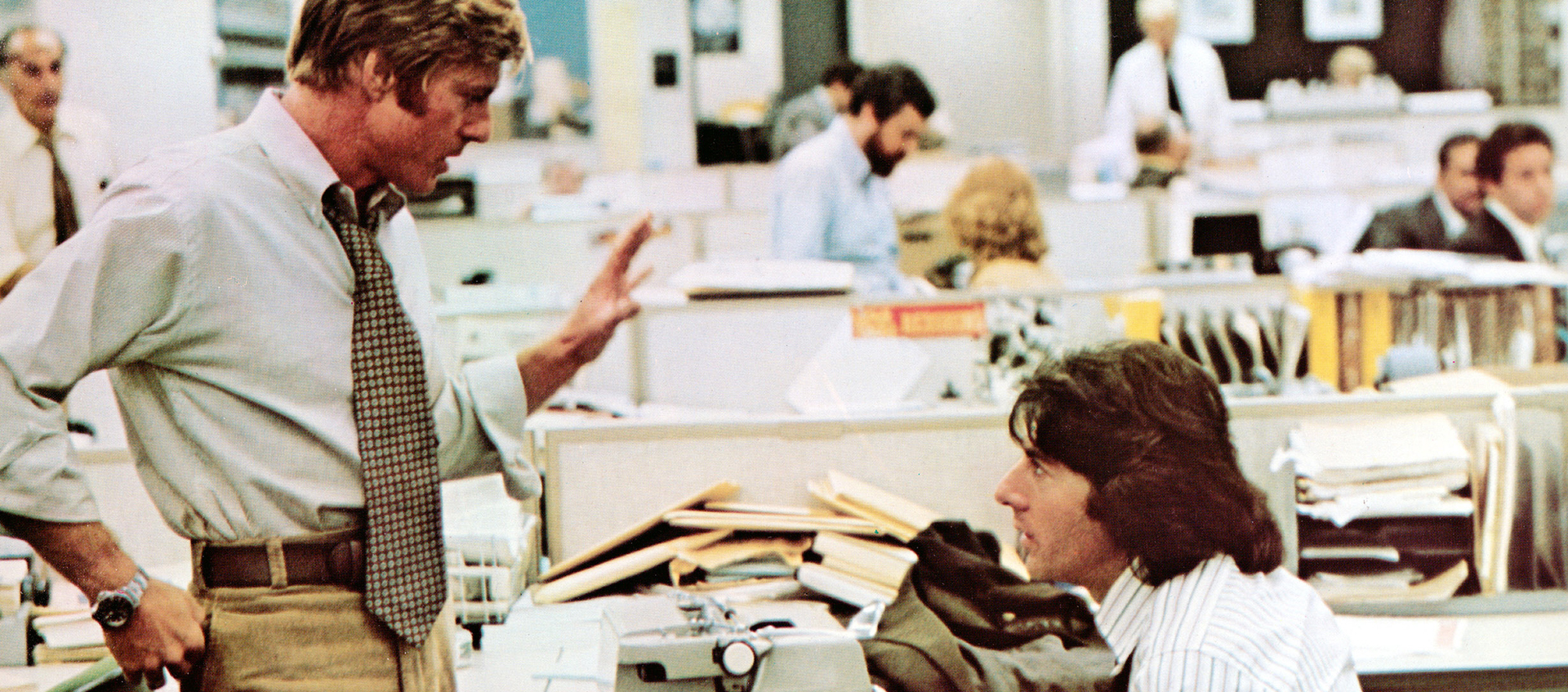 Two men are talking in a busy newsroom, one of them is standing and gesturing while the other sits. 