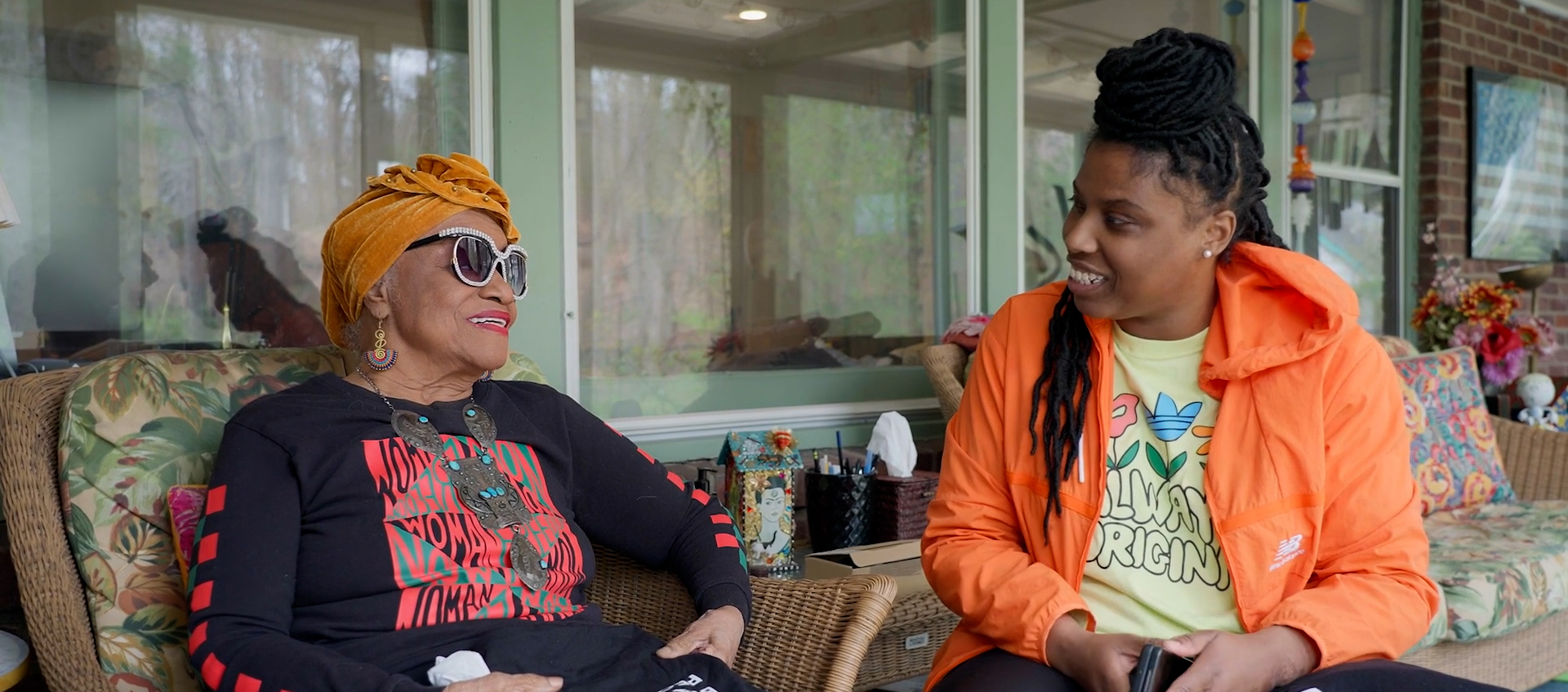 Two Black women are sitting on a porch talking to each other; one is older wearing sunglasses and a head scarf, the other is wearing a jacket. 