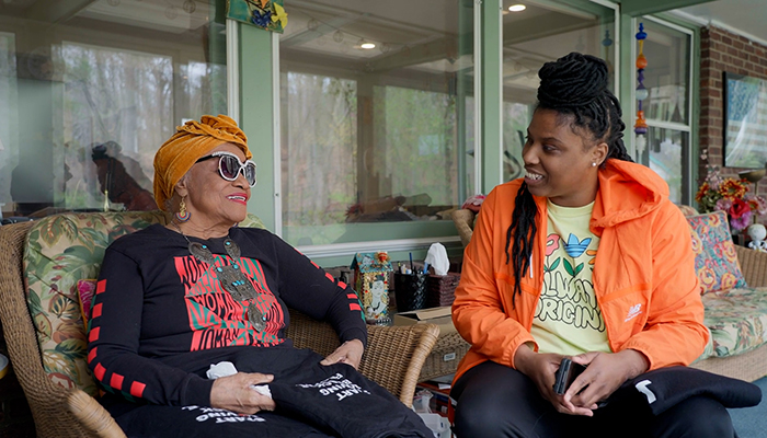 Two Black women are sitting on a porch talking to each other; one is older wearing sunglasses and a head scarf, the other is wearing a jacket. 