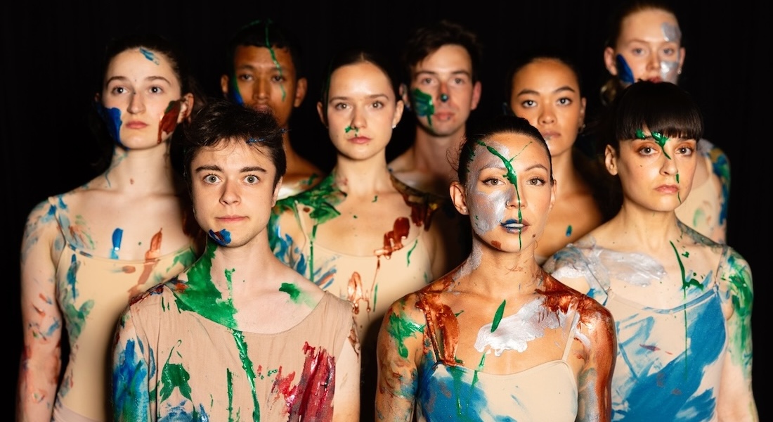 A group of nine dancers in neutral leotards stands together, covered in paint, in front of a black background