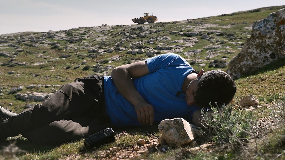 A young man lies on his side in a rocky, grassy field with a camera by his side.