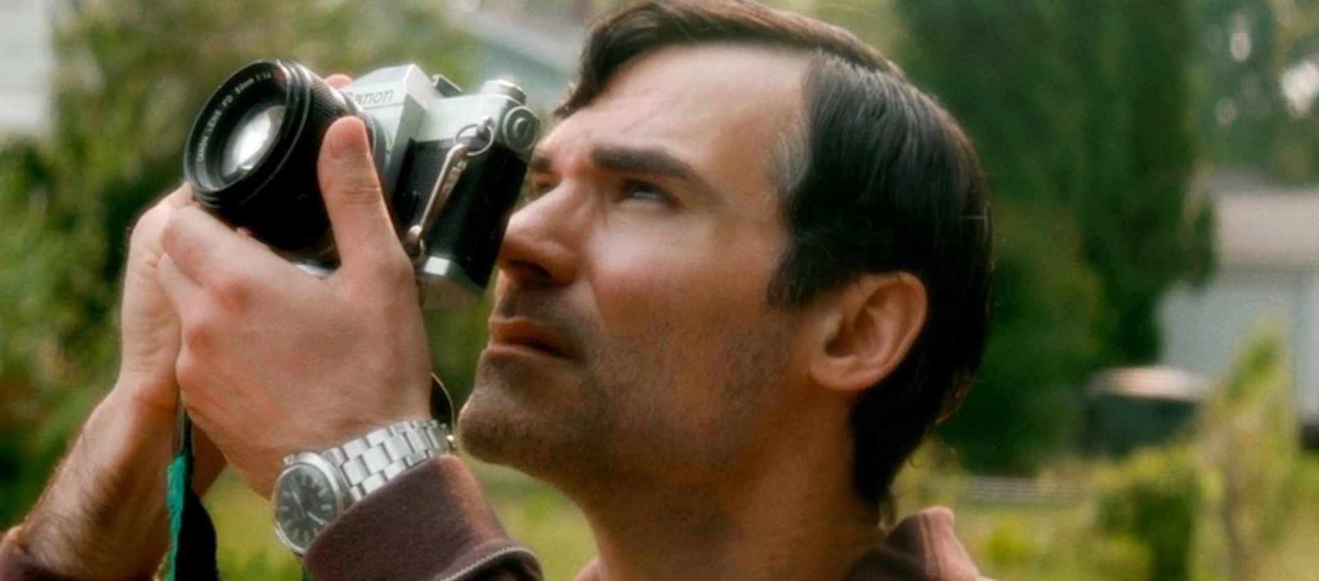 A man with short, dark hair stands outside and looks through the viewfinder of a 35mm camera.