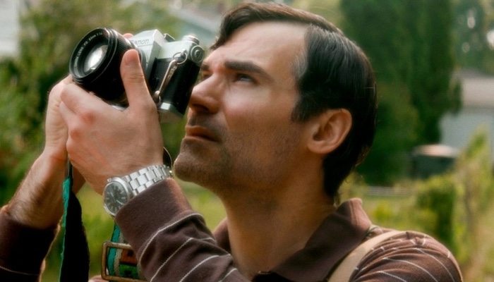 A man with short, dark hair stands outside and looks through the viewfinder of a 35mm camera.