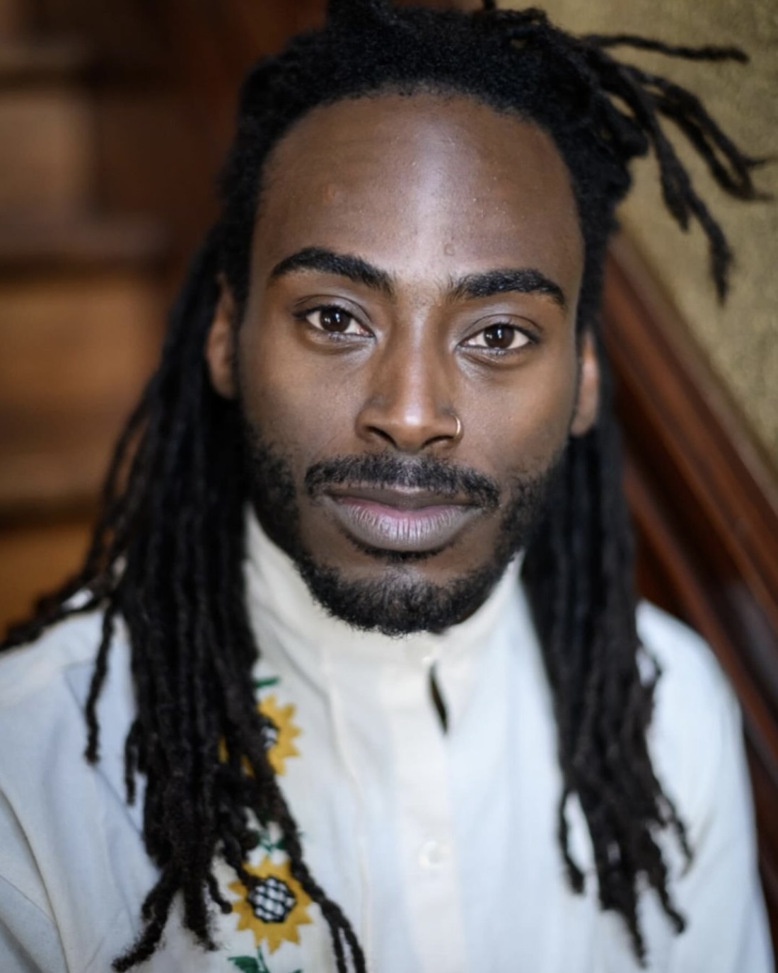 A man with long dreads, wearing a white shirt with floral embroidery, smiles for the camera.