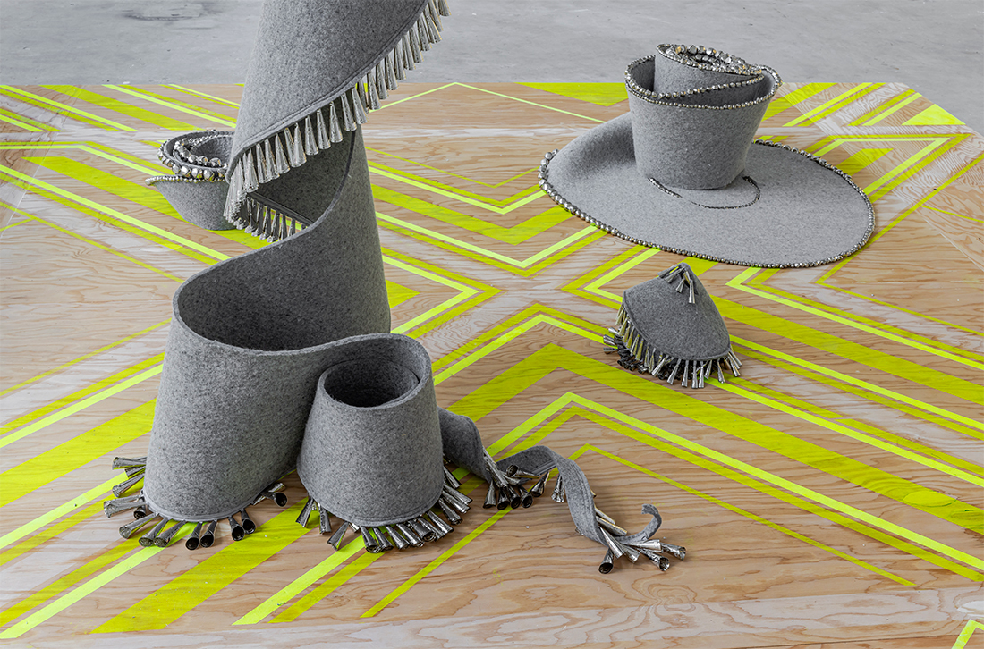 Closeup of a hanging spiral shaped length of gray felt edged with tin jingles that drapes onto a patterned plinth supporting three other felt pieces.
