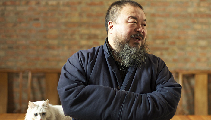 Photo of Ai Weiwei, a man with a beard wearing a blue jacket with his arms folded standing in front of a brick wall.