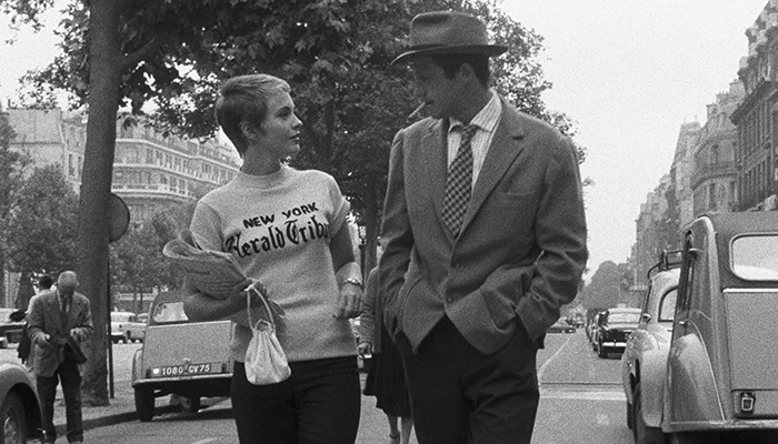 A man and a woman walking down the middle of a busy street filled with cars and lined by trees. 