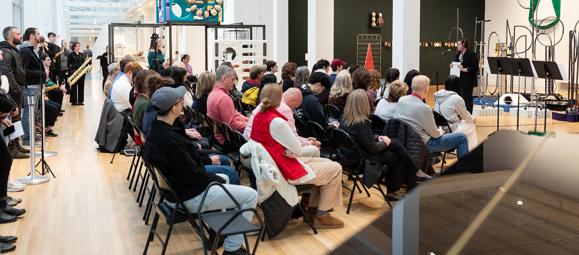 A seated and standing audience watches a musician who stands in front of sculptures in a large gallery space. 