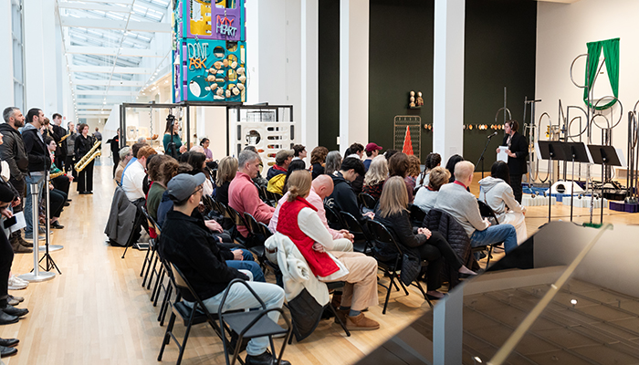 A seated and standing audience watches a musician who stands in front of sculptures in a large gallery space. 