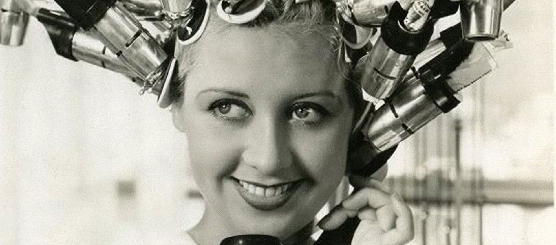 Black and white image of a smiling woman with her hair up in a large hair curling machine.