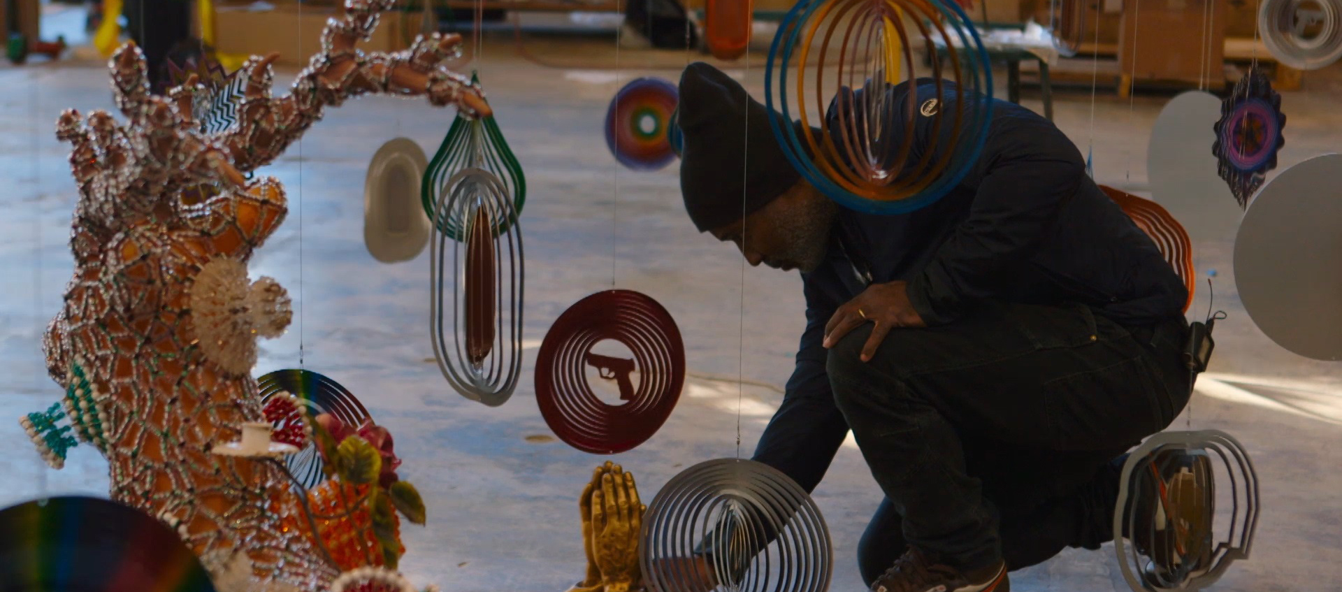 A man kneels on the floor surrounded by colorful hanging sculptures.