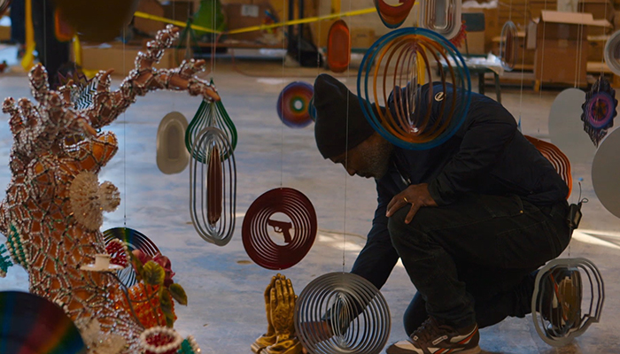 A man kneels on the floor surrounded by colorful hanging sculptures.