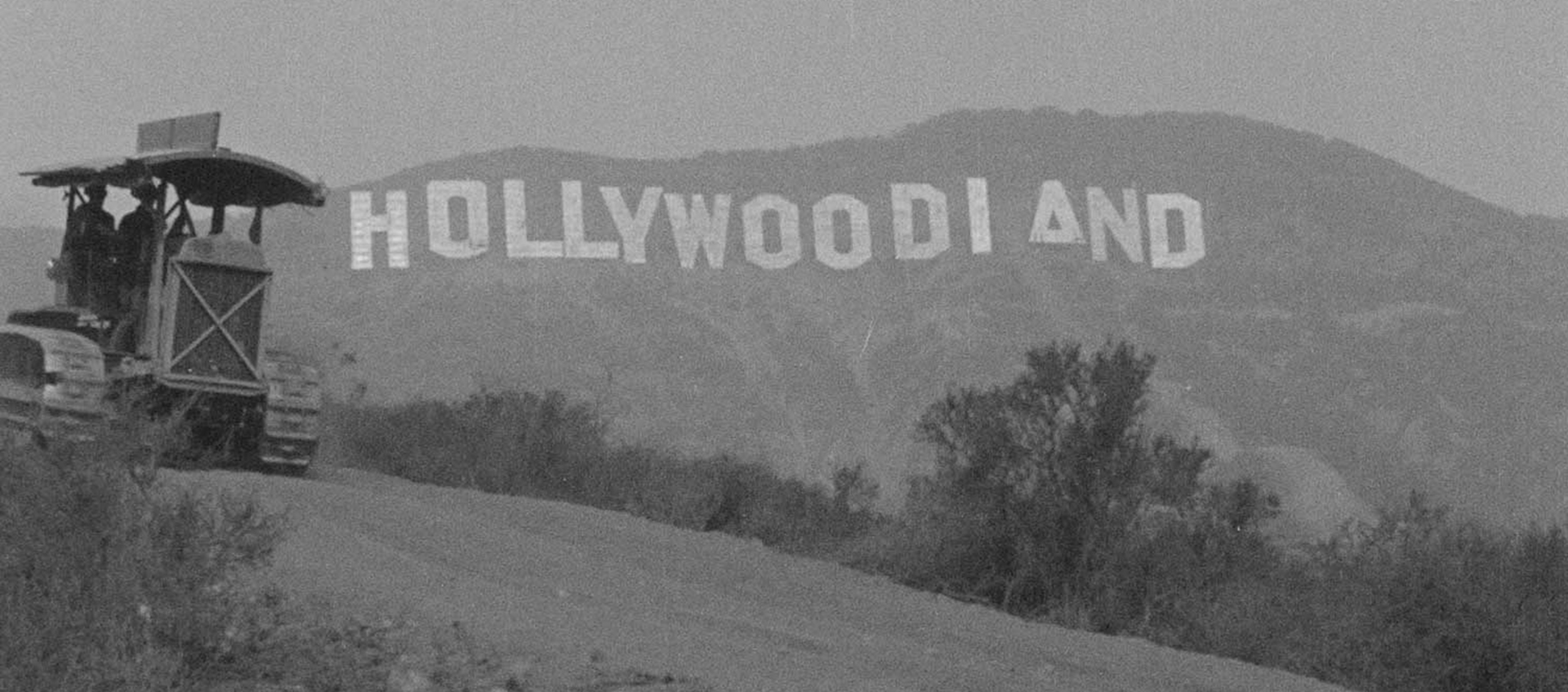 Black and white image of the original Hollywoodland sign on a hillside, partially obscured by a hill and bushes, with a vehicle nearby.