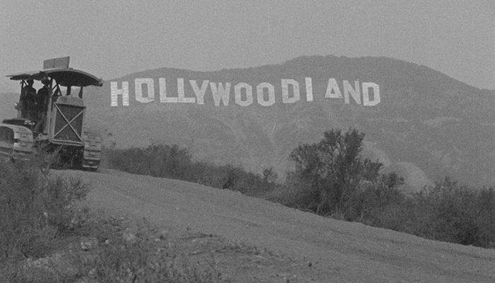 Black and white image of the original Hollywoodland sign on a hillside, partially obscured by a hill and bushes, with a vehicle nearby.